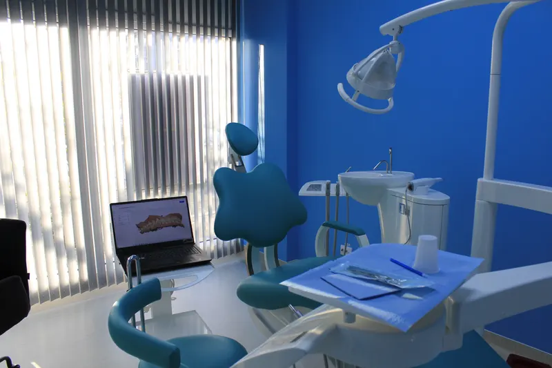 Unit dentar turcoaz lângă fereastra cu jaluzele verticale, cu un laptop pe masa de lucru