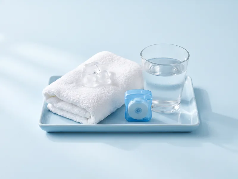Prosop alb cu cuburi de gheață, pahar cu apă călduță și ață dentară pe o tavă albastru pal